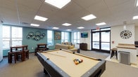 This community room is a well-appointed, single-floor gathering space designed to encourage socializing and recreation. At the heart of the room sits a full-size billiards table, making it an immediate focal point for entertainment. The space features a relaxed lounge area with comfortable seating arranged around a wall-mounted flat-screen television, ideal for casual gatherings or watching a favorite show. A foosball table adds another layer of recreational activity, rounding out the game-room atmosphere. Large windows along the perimeter invite abundant natural light, creating a bright and welcoming environment throughout the day. A set of glass double doors provides easy access and adds to the open, airy feel of the room. The drop ceiling is fitted with recessed panel lighting that ensures consistent, even illumination. Accent walls in a calming teal tone, complemented by decorative wall art and metallic candle ring fixtures, add personality and visual interest to the space. The combination of carpeted flooring, thoughtful décor, and versatile amenities makes this community room a comfortable and functional hub for residents to connect and unwind.