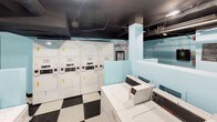 Located in Dallas, this shared laundry facility is a practical and functional space designed to accommodate multiple users efficiently. The room is defined by its distinctive black-and-white checkered tile flooring, which adds a classic visual character to the utilitarian environment. Light blue painted walls provide a clean, bright contrast against the exposed black ceiling, through which ductwork, piping, and overhead lighting are clearly visible, lending the space an industrial aesthetic.

The facility is well-stocked with a generous lineup of commercial-grade top-loading washing machines and stacked dryer units, all finished in white and arranged to maximize the use of available floor space. Folding baskets and a wooden folding surface are conveniently positioned for users. Instructional signage for washing and drying procedures is posted on the walls, ensuring ease of use.

A separate alcove area houses a bank of storage lockers, offering secure personal storage for residents. A wooden bench is positioned nearby, providing a comfortable place to wait. A frosted-glass interior door connects the main laundry area to an adjacent space, enhancing flow through the facility. Overall, the space is clean, organized, and thoughtfully laid out to serve as a reliable shared amenity.