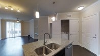 This single-story space showcases an open-concept design that connects the kitchen and living areas into one cohesive living environment. The kitchen serves as a central hub, anchored by a large island topped with speckled granite countertops and fitted with a double stainless steel undermount sink and a sleek pull-down faucet. Two cylinder pendant lights hang above the island, providing focused task lighting with a contemporary aesthetic. Dark cabinetry on the island contrasts with the neutral warm-beige walls and is complemented by stainless steel appliances, including a dishwasher. Crown molding runs along the upper perimeter of the space, adding a refined architectural detail throughout. Dark hardwood flooring extends across the main living area, grounding the space with a rich, polished look. A dedicated laundry alcove is neatly integrated into the layout, housing a side-by-side washer and dryer behind bi-fold doors for convenient in-unit access. The open sightlines from the kitchen into the living area enhance the sense of spaciousness, while recessed lighting supplements the pendant fixtures for consistent, well-distributed illumination throughout.