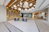Located at 101 SW 1st St in Dania Beach, Florida, this single-floor space spans a generous footprint and delivers a striking first impression through its thoughtfully curated modern design. The grand lobby serves as the centerpiece, featuring polished terrazzo flooring with inlaid accent banding, soaring ceilings adorned with warm wood slat paneling, and a dramatic cluster of globe pendant lights that create an elegant, sculptural focal point overhead. Backlit vertical wood fin walls cast a warm ambient glow, while floor-to-ceiling glass walls and expansive glazed entry doors flood the interior with abundant natural light and offer views of the surrounding palm-lined streetscape. A custom live-edge communal table with bar-height stools anchors the lobby, complemented by stylish lounge seating areas with patterned area rugs and warm-toned sofas. Sheer drapery panels soften the glass perimeter, adding a refined residential quality to the commercial setting. Three private glass-enclosed offices provide focused work areas, each finished with patterned carpet, built-in wood millwork, and modern ergonomic furnishings. A dedicated mail room features a full bank of resident mailboxes framed by a dark accent wall and warm wood trim. Wide, well-lit corridors with terrazzo flooring and textured wall treatments connect all areas seamlessly throughout the space.
