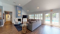 This single-story space showcases a cohesive coastal theme throughout its open, well-connected layout. The living area immediately draws attention with its soaring vaulted ceilings that amplify the sense of openness, complemented by recessed lighting and a striking blue accent wall that anchors the room. A white brick fireplace serves as a focal point, while large windows fitted with plantation shutters allow generous natural light to filter in and offer views of an outdoor pool area. A glass-top side table resting on a natural driftwood base reinforces the nautical aesthetic alongside beach-themed artwork and décor.

Rich, dark hardwood-style flooring flows continuously through the living and dining areas, creating visual cohesion. The dining area features a round glass-top table with woven chairs and transitions seamlessly into a built-in counter workspace equipped with bar-height stools — a functional and stylish detail. Coastal wall accents, including a large decorative clock and porthole-style mirrors, enhance the thematic continuity.

The kitchen is outfitted with white shaker-style cabinetry, granite countertops, stainless steel appliances, and earthy slate tile flooring, offering a practical contrast to the warmer tones of the main living space. Together, the rooms create a relaxed, resort-inspired ambiance throughout.