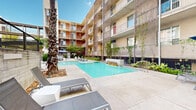 This outdoor courtyard amenity space is centrally situated within a contemporary multi-story residential complex, offering a well-appointed resort-style experience. The centerpiece is a generously sized swimming pool with clear, blue-tinted water, complemented by a built-in water feature that adds both visual appeal and ambient sound to the environment.

The pool deck is finished with large-format concrete pavers, providing ample space for relaxation. Modern lounge chairs and side tables are thoughtfully arranged around the pool, offering residents comfortable spots to unwind. Shaded seating areas with large umbrellas and upholstered outdoor furniture provide additional gathering spaces throughout the courtyard.

The surrounding architecture features a bold, modern aesthetic with colorful accent panels in warm red and neutral tones, exposed concrete balconies, and open-air corridors across multiple floors. Metal trellis screens along the building facades support climbing vines, softening the structure and adding a natural, green element to the urban setting.

Landscaping throughout the courtyard includes tall palm trees, flowering shrubs such as blue plumbago, ornamental grasses, and layered plantings along the perimeter. Together, these elements create a lush, resort-like atmosphere that balances architectural structure with organic greenery, making this courtyard a standout communal amenity within the complex.