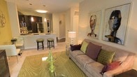 This single-floor residence showcases an open-concept layout that integrates the living and kitchen areas into one cohesive, flowing space. The living room is anchored by a plush taupe sofa dressed with decorative pillows in deep purple and green, set atop a bold patterned area rug in complementary green tones. A glass-top accent table adds a modern touch to the seating area, while framed artwork on the warm beige walls contributes a curated, polished aesthetic.

The kitchen is positioned directly adjacent to the living space, separated by a peninsula-style counter with bar seating accommodating two stools. Dark cabinetry provides a striking contrast against the light countertops, while stainless steel appliances, including a refrigerator, lend a sleek, contemporary finish. Pendant lighting above the counter and a flush ceiling fixture illuminate the kitchen with a warm glow, enhancing the inviting atmosphere.

A corridor visible from the main living area suggests additional rooms beyond, including what appears to be laundry facilities. The overall ambiance is warm, modern, and well-appointed, creating a comfortable and stylish everyday living environment.