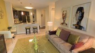 This single-floor residence showcases an open-concept layout that integrates the living and kitchen areas into one cohesive, flowing space. The living room is anchored by a plush taupe sofa dressed with decorative pillows in deep purple and green, set atop a bold patterned area rug in complementary green tones. A glass-top accent table adds a modern touch to the seating area, while framed artwork on the warm beige walls contributes a curated, polished aesthetic.

The kitchen is positioned directly adjacent to the living space, separated by a peninsula-style counter with bar seating accommodating two stools. Dark cabinetry provides a striking contrast against the light countertops, while stainless steel appliances, including a refrigerator, lend a sleek, contemporary finish. Pendant lighting above the counter and a flush ceiling fixture illuminate the kitchen with a warm glow, enhancing the inviting atmosphere.

A corridor visible from the main living area suggests additional rooms beyond, including what appears to be laundry facilities. The overall ambiance is warm, modern, and well-appointed, creating a comfortable and stylish everyday living environment.