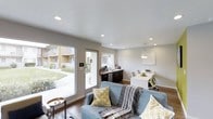 This single-floor space offers a thoughtfully designed open-concept layout that seamlessly integrates a living area, kitchenette, and dining zone into one cohesive environment. The living area is anchored by a blue sofa accented with yellow and neutral throw pillows, complemented by warm hardwood-style flooring that runs throughout the space.

Large glass doors along one wall flood the interior with abundant natural light while providing direct views of a landscaped outdoor area, creating a welcoming indoor-outdoor connection. Recessed ceiling lighting supplements the natural light, ensuring the space remains bright throughout the day.

The compact kitchenette features dark cabinetry, a sink, and countertop appliances, offering functional utility without occupying significant floor space. Adjacent to the kitchenette, a sleek white dining table paired with modern chairs serves as a dedicated workspace or dining area, illuminated by a contemporary pendant light fixture above.

The neutral gray wall tones and clean white ceilings create a calm, modern aesthetic throughout, while the efficient layout maximizes usability across the open floor plan.