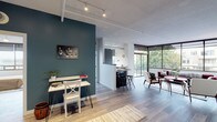Located at 1224 West Riverside Avenue in downtown Spokane, this modern single-floor unit blends contemporary design with urban convenience. The open-concept living and dining area is bathed in natural light through expansive floor-to-ceiling windows, offering sweeping views of the Spokane cityscape. Track lighting and exposed ductwork lend an industrial-chic character, while light wood-look flooring flows throughout. A bold teal accent wall anchors the living space, which features a wall-mounted electric fireplace as a stylish focal point. The sleek kitchen is outfitted with high-gloss cabinetry, stainless steel appliances, white countertops, and a striking wraparound window that frames panoramic city views — an exceptional feature for cooking and entertaining. A dedicated workspace nook adds functional versatility. The unit offers two bedrooms, including one with large windows overlooking the city and a frosted-glass sliding barn door leading to an en suite bathroom. The en suite features a glass-enclosed shower, gray vanity, and mosaic tile backsplash. A second bathroom includes a tiled tub-shower combo. A sunlit sitting nook with additional city views completes this well-appointed urban retreat.