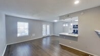 This contemporary apartment showcases a spacious, open-concept design with a seamless flow between living and dining areas. The interior boasts sleek wood-look flooring throughout, complemented by light gray walls and recessed lighting. The updated kitchen features white cabinetry, pendant lighting, and black appliances, including a large refrigerator. A cozy fireplace adds warmth to the living space. The bathroom offers a modern vanity with ample storage. French doors and large windows provide natural light, while the community exterior features well-maintained landscaping and wooden fencing, creating a welcoming atmosphere.