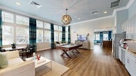 This single-story community clubhouse is thoughtfully designed with a cohesive coastal-inspired color palette of teal, blue, and neutral tones, unified by warm hardwood-style flooring throughout. The spacious main lounge area serves as the centerpiece of the space, featuring a statement pool table beneath a sculptural gold orb chandelier, complemented by comfortable seating, a flat-screen television atop a textured media console, and floor-to-ceiling windows dressed in bold teal and navy striped drapery that flood the room with natural light. An intimate seating nook adjacent to the lounge offers a round dining table and mixed accent chairs, surrounded by oversized windows that frame outdoor views. A vibrant reception and leasing area showcases deep blue accent walls, pendant lighting, a built-in counter-height workstation, and welcoming teal upholstered chairs. The kitchenette is well-appointed with gray shaker-style cabinetry extending to the ceiling, granite countertops, a white tile backsplash, a stainless steel refrigerator, and a full sink setup. A separate lounge zone with bold blue walls, patterned valances, and glass-paneled French doors provides additional gathering space, completing this well-appointed and visually cohesive community facility.