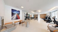 Located at 22 Hubbard Road in Wilton, Connecticut, this modern fitness center offers a well-appointed, single-floor layout designed to support a comprehensive workout experience. The space is divided into two functional zones that together provide a full range of exercise options.

The main workout area is bright and open, featuring light-toned flooring and white walls that create a clean, energizing atmosphere. A row of large windows along one wall floods the cardio section with natural light, where treadmills, elliptical trainers, and stationary bikes are neatly arranged. Overhead linear lighting supplements the natural illumination throughout.

The strength training section is anchored by a warm wood-paneled accent wall, adding a refined contrast to the otherwise contemporary design. Full-length mirrors amplify the sense of space and assist with form during workouts. The area is outfitted with a cable resistance machine, a dumbbell rack, adjustable benches, and a variety of stability balls and resistance equipment organized along the mirrored wall. A bold photographic art piece adds a dynamic visual element to the space.

A welcoming lounge area near the entrance features casual seating with contemporary chairs and a round side table, providing a space to rest or consult. A water fountain is also conveniently located near the entry. The ceiling-mounted HVAC system ensures year-round climate comfort throughout the facility.