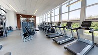 This fitness center is designed to provide a comprehensive and energizing workout environment. The space is defined by an expansive wall of floor-to-ceiling windows that floods the interior with natural light and offers unobstructed views of the surrounding greenery and outdoor infrastructure, creating an open and motivating atmosphere for exercise.

The cardio zone is prominently featured along the window wall, housing a row of modern treadmills and elliptical cross-trainers, all positioned to allow users to enjoy the outdoor scenery during their workouts. Adjacent to the cardio equipment, a dedicated strength training area includes a selection of selectorized resistance machines and weight benches, catering to a full range of muscle groups.

The flooring throughout consists of durable, commercial-grade rubber material designed to withstand heavy use while providing comfort and safety. The ceiling is finished with a drop-tile system complemented by recessed and linear lighting, ensuring consistent illumination across the entire floor.

An exposed brick accent wall adds a touch of architectural character, contrasting with the otherwise sleek and contemporary design. Mirrors are strategically placed to enhance the sense of space and allow users to monitor their form. Overall, the fitness center balances functionality with a bright, welcoming ambiance.