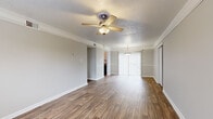 This single-story residence presents a clean, neutral interior designed for comfortable living. The main living area is generously proportioned, featuring warm wood-look luxury vinyl flooring that extends throughout the common spaces. Crown molding lines the ceilings, adding a refined architectural detail, while a ceiling fan with a light fixture provides both air circulation and ambient lighting. A sliding glass door at the far end of the living area allows natural light to filter in, creating a bright and open atmosphere.

Adjacent to the living space is a dining area, distinguished by its own pendant light fixture and a chair-rail accent along the walls, with a sliding door leading to a connecting room. The kitchen is outfitted with wood-look flooring and includes a stainless steel refrigerator.

The bedrooms are finished with plush neutral carpeting and each features a ceiling fan and natural light from windows overlooking greenery. One bedroom notably includes dual sliding closets, providing substantial storage capacity.

The bathroom is thoughtfully appointed with wood-look flooring, a tub-shower combination with white tile surround, and accessibility grab bars, complemented by a vanity with granite-style countertops. Throughout, the neutral gray walls and white trim create a cohesive, move-in-ready aesthetic.