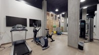 This well-appointed fitness center offers a thoughtfully designed workout environment that combines industrial aesthetics with practical functionality. The space features striking exposed concrete columns and dark ceilings that contrast beautifully with the white walls and light wood-toned flooring, creating a modern and energizing atmosphere.

The cardio zone is equipped with a treadmill, elliptical trainer, and stationary bike, all positioned alongside wall-mounted flat-screen televisions for entertainment during workouts. Toward the rear of the space, a dedicated functional training area houses a variety of exercise tools, including medicine balls, balance balls, and weight plates neatly organized on a rack. A large floor-to-ceiling mirror along one wall enhances the sense of openness while allowing users to monitor their form.

Warm wood slatted partitions add a refined architectural touch, softening the industrial elements and defining distinct zones within the space. Overhead industrial-style lighting ensures the entire area is well illuminated. The combination of cardio machines, functional fitness equipment, and a clean, open layout makes this fitness center a comprehensive and inviting facility suited for a wide range of workout routines.