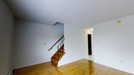 This well-appointed multi-level space is spread across three floors, offering a versatile and functional layout with a classic design aesthetic. The main level immediately sets the tone with warm, honey-toned hardwood floors that extend throughout, reflecting light and adding a sense of warmth and continuity to the space. Crisp white walls provide a clean, neutral backdrop that enhances the brightness of the interior. A standout architectural feature is the wood staircase accented with a sleek black iron railing, which serves as both a functional connector between floors and a subtle design focal point. The staircase rises from the main living area, hinting at the additional space available on the upper levels. A wall-mounted light fixture adds ambient illumination, while an open doorway leads to an adjacent room or hallway, suggesting a thoughtful and connected floor plan. The overall ambiance is understated and inviting, with a timeless quality suited to a variety of uses. The three-floor configuration provides ample vertical space, making this property ideal for those seeking separation between living, working, and private areas across distinct levels.