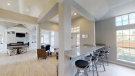 This single-floor community clubhouse is designed with an open-concept layout that seamlessly connects multiple functional areas, all finished with light hardwood flooring and a cohesive neutral gray-and-white palette. Soaring vaulted and beamed ceilings throughout the main gathering spaces create a bright, airy atmosphere, further enhanced by abundant natural light streaming through large multi-pane windows.

The central lounge area serves as the heart of the space, anchored by a dramatic floor-to-ceiling marble-clad fireplace with a wall-mounted flat-screen television above. Comfortable dark leather seating arrangements invite relaxation. Adjacent to the lounge, a granite-topped bar counter with modern swivel barstools provides an ideal spot for casual socializing, with views into connected activity areas.

The game room, visible from the main gathering space, features a pool table and additional seating, while a dedicated dining area offers a formal table with upholstered leather chairs beneath a tray ceiling with crown molding and garden-facing windows. A fitness center, accessible through a glass-paned door, is equipped with cardio machines. The overall design balances contemporary finishes with welcoming architectural details, making this a versatile and polished communal facility.