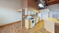 This single-floor loft space masterfully blends industrial character with functional living. The open main area is defined by striking exposed brick walls, soaring ceilings with visible wooden beam construction, and industrial ductwork running overhead — all hallmarks of a thoughtfully preserved building conversion. Ceiling fans are strategically placed throughout, and a row of large windows along the brick wall floods the space with abundant natural light while offering views of the surrounding area. Rich hardwood flooring extends across the open living area, complementing the warm tones of the brick and wood overhead. A freestanding electric fireplace adds a cozy focal point to the expansive main room. The kitchen is well-appointed with light wood cabinetry, a full suite of white appliances including a range, microwave, dishwasher, and refrigerator, and generous counter space. A large kitchen island with pendant lighting above anchors the kitchen area and provides additional workspace and seating potential. The bathroom is clean and practical, featuring a single-sink vanity with wood cabinetry, tile flooring, and a walk-in shower stall. The overall ambiance of the space is open, airy, and rich with architectural character.