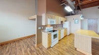 This single-floor loft space masterfully blends industrial character with functional living. The open main area is defined by striking exposed brick walls, soaring ceilings with visible wooden beam construction, and industrial ductwork running overhead — all hallmarks of a thoughtfully preserved building conversion. Ceiling fans are strategically placed throughout, and a row of large windows along the brick wall floods the space with abundant natural light while offering views of the surrounding area. Rich hardwood flooring extends across the open living area, complementing the warm tones of the brick and wood overhead. A freestanding electric fireplace adds a cozy focal point to the expansive main room. The kitchen is well-appointed with light wood cabinetry, a full suite of white appliances including a range, microwave, dishwasher, and refrigerator, and generous counter space. A large kitchen island with pendant lighting above anchors the kitchen area and provides additional workspace and seating potential. The bathroom is clean and practical, featuring a single-sink vanity with wood cabinetry, tile flooring, and a walk-in shower stall. The overall ambiance of the space is open, airy, and rich with architectural character.