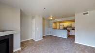 This single-floor residence offers a well-organized, functional layout designed for comfortable everyday living. The open-concept living area features neutral gray walls, plush wall-to-wall carpeting, and wood-look vinyl flooring that transitions seamlessly into the kitchen. Built-in open shelving adds a practical decorative touch to the living space, while a flush-mount ceiling fixture provides warm ambient lighting.

The kitchen is efficiently designed with white cabinetry, dark laminate countertops, and a full suite of white appliances including a refrigerator, electric range, and over-the-range microwave. The kitchen opens directly to the living area via a raised bar-height peninsula, creating a connected, social atmosphere. Wood-look flooring runs throughout the kitchen for easy maintenance.

The bedroom is generously proportioned with soft carpeting underfoot, a tray ceiling detail with a ceiling fan fixture, and a sliding glass door that opens directly onto a private balcony — bringing in natural light and outdoor views. The bedroom is further complemented by a walk-in closet with built-in shelving and rod storage, providing ample organization space.

The bathroom features wood-look flooring, a large mirror, and clean, simple fixtures. Throughout the space, a consistent soft gray color palette and white trim create a calm, cohesive aesthetic.