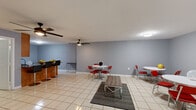 This single-floor space is thoughtfully laid out to maximize functionality across its rooms. The open living area is finished with large-format light ceramic tile flooring that runs consistently throughout, lending a clean and cohesive look. Gray-painted walls provide a neutral backdrop, while multiple ceiling fans with integrated lighting keep the space comfortable and well-lit. The living area accommodates multiple retro-style round dining tables with chrome-framed red chairs, creating a casual, communal atmosphere, complemented by a wall-mounted television.

The dual kitchen areas are a standout feature, each equipped with warm-toned wood cabinetry, dark granite-style countertops, and black appliances including a refrigerator and over-the-range microwave. A central kitchen island with a built-in sink adds practical counter space and further defines the kitchen zone from the open living area.

The bathroom is clean and straightforward, featuring the same light tile flooring, a wood-vanity with sink and mirror, and a toilet — all set against the consistent gray wall color. The overall ambiance of the space is functional and social, well-suited for communal or multi-purpose use.