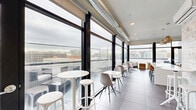 This contemporary rooftop amenity space offers an elevated urban experience, combining an interior lounge with an expansive open-air terrace. The interior is defined by dramatic floor-to-ceiling black-framed windows that flood the space with natural light and frame sweeping panoramic city views. Large-format light grey tiles run throughout the interior, complementing the clean, white walls and exposed metallic spiral ductwork overhead, lending an industrial-chic aesthetic to the design.

The interior lounge area features a well-appointed kitchenette with a large white island, warm wood cabinetry, vibrant blue tile backsplash, and pendant lighting, making it well-suited for gatherings and events. A mix of high-top bistro tables, modern seating, and casual lounge chairs are arranged throughout, providing flexible configurations for socializing or working.

A glass-and-black-framed door leads to the open-air terrace, where glass panel railings preserve unobstructed sightlines across the city skyline. The terrace is furnished with concrete lounge chairs, cushioned seating, and high stools, all set against a textured brick accent wall. The reflective terrace flooring and expansive sky views create a striking outdoor setting. Together, the interior and exterior areas offer a cohesive, sophisticated rooftop experience with a distinctly modern urban character.