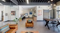 Located at 2424 N Tustin Ave in Santa Ana, California, this single-story community space is designed with a contemporary, open-concept layout that seamlessly blends functionality with modern aesthetics. Exposed dark steel ceiling beams contrast against crisp white walls, lending an industrial-chic character throughout the space, while polished concrete flooring ties each area together cohesively.

The main lounge area is generously proportioned and anchored by a collection of cognac leather sofas and armchairs accented with vibrant, striped throw pillows. A large communal wood-top dining table with sculptural metal legs and industrial pendant lighting serves as a central gathering point, ideal for both casual meetings and collaborative work sessions. A wall-mounted flat-screen TV adds an entertainment element to the lounge.

A glass-walled partition elegantly separates the leasing or administrative office, which is equipped with multiple computer workstations and ample natural light. This transparency maintains an open, connected atmosphere throughout the interior. A dedicated computer workstation nook along one wall provides additional workspace for residents.

Floor-to-ceiling glass doors open directly to an outdoor patio, where umbrella-shaded seating areas are visible, flooding the interior with abundant natural light and creating a strong indoor-outdoor connection. Colorful artwork adorns the walls, and lush potted plants are positioned throughout, adding warmth and vibrancy to the overall ambiance.