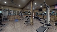 Located in Spring Hill, Tennessee, this fitness facility combines rustic charm with modern functionality, creating an inviting and well-appointed space for exercise and wellness. The single-floor layout is generously sized, offering ample room for members to move freely between stations and equipment zones.

The architectural design is a standout feature, showcasing exposed heavy timber wood beam columns set atop stacked natural stone bases that run throughout the floor. Matching stone accent walls add warmth and character, while the neutral-toned walls and recessed lighting create a welcoming, well-lit environment. Ceiling fans are positioned throughout to aid in air circulation.

The facility is equipped with an extensive array of commercial-grade machines, including a variety of Life Fitness and cable-based strength machines, a functional trainer, adjustable benches, a dumbbell rack, and multiple cardio options such as treadmills, stationary bikes, and a rowing machine. Floor space is also available for stretching and mat-based exercises.

Large mirrors line the walls, enhancing the sense of openness and allowing members to monitor their form during workouts. Multiple wall-mounted flat-screen televisions are positioned throughout the space for entertainment. Wood-look flooring ties the entire aesthetic together, reinforcing the facility's warm, lodge-inspired atmosphere.