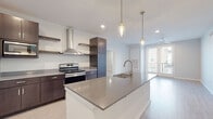 This contemporary single-floor unit showcases a thoughtfully designed open-concept layout with clean lines and modern finishes throughout. The kitchen serves as the centerpiece, featuring a large island with an integrated sink, sleek quartz countertops, dark espresso cabinetry, stainless steel appliances including an electric range and built-in microwave, a wall-mounted range hood, and open floating shelves. Pendant lighting above the island adds a refined touch, while light wood-look flooring flows seamlessly into the adjacent living area, which is equipped with a ceiling fan and access to a private balcony. The bedrooms are bright and well-proportioned, each with ceiling fans and natural light from windows with blinds. Storage is a standout feature, with generously sized walk-in closets outfitted with extensive wire shelving systems. The bathrooms are elegantly appointed with dark espresso vanities, gray quartz countertops, and illuminated mirrors. The primary bath includes dual vanities and a separate tub/shower combination. A secondary kitchenette area with upper and lower cabinetry, a full-size refrigerator, and a microwave provides added flexibility. The consistent use of light flooring, neutral wall tones, and recessed lighting gives the entire space a bright, cohesive, and modern ambiance.