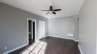 This single-story space is defined by its clean, understated design and practical layout. The room features rich, dark hardwood flooring that contrasts sharply with the light gray walls, creating a balanced and neutral aesthetic throughout. Crown molding runs along the ceiling perimeter, adding a subtle architectural detail that lends a polished finish to the space.

A ceiling fan with a built-in light fixture is centrally mounted, providing both air circulation and ambient lighting. A wall-mounted heater unit is positioned along one wall, offering a supplemental heating solution. White baseboards and door trim provide clean contrast against the gray walls, reinforcing a cohesive color palette.

Natural light filters into the room through a window, casting a grid-patterned shadow across the hardwood floors and enhancing the sense of openness. Two white-framed doorways are visible — one leading to what appears to be a closet or storage area, and another opening to an adjoining room. The overall ambiance is calm and functional, with a neutral palette that offers a versatile foundation for a variety of uses or furnishing styles.