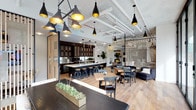The lobby at 1810 State Street in San Diego presents a thoughtfully designed, open-plan environment that seamlessly integrates multiple functional areas into one cohesive space. The single-floor layout features an exposed ceiling with visible ductwork and piping painted white, lending an industrial character that is softened by warm, contemporary finishes throughout.

A striking collection of black pendant lights with gold interiors hangs at varying heights, creating a dramatic focal point overhead while casting a warm, inviting glow across the space. The light wood flooring runs throughout, unifying the distinct zones and contributing to an airy, welcoming atmosphere.

A full kitchen anchors one side of the space, featuring dark cabinetry, a white countertop island, and bar-height seating with black stools — ideal for casual gatherings or collaborative meetings. Adjacent dining and lounge areas offer a variety of seating arrangements, including wooden tables with mixed chairs and a comfortable navy sofa, providing flexible options for residents or visitors.

A slatted wood partition and a vertical rod room divider subtly define zones without sacrificing the open feel. Touches of greenery, including tabletop planters and potted plants, add life and freshness to this well-appointed, multifunctional lobby.