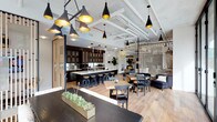 The lobby at 1810 State Street in San Diego presents a thoughtfully designed, open-plan environment that seamlessly integrates multiple functional areas into one cohesive space. The single-floor layout features an exposed ceiling with visible ductwork and piping painted white, lending an industrial character that is softened by warm, contemporary finishes throughout.

A striking collection of black pendant lights with gold interiors hangs at varying heights, creating a dramatic focal point overhead while casting a warm, inviting glow across the space. The light wood flooring runs throughout, unifying the distinct zones and contributing to an airy, welcoming atmosphere.

A full kitchen anchors one side of the space, featuring dark cabinetry, a white countertop island, and bar-height seating with black stools — ideal for casual gatherings or collaborative meetings. Adjacent dining and lounge areas offer a variety of seating arrangements, including wooden tables with mixed chairs and a comfortable navy sofa, providing flexible options for residents or visitors.

A slatted wood partition and a vertical rod room divider subtly define zones without sacrificing the open feel. Touches of greenery, including tabletop planters and potted plants, add life and freshness to this well-appointed, multifunctional lobby.