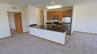 This single-floor space showcases a thoughtfully designed open-concept layout that seamlessly integrates the kitchen and living areas. The kitchen is a standout feature, equipped with a full suite of stainless steel appliances, including a refrigerator, range with oven, and built-in microwave. Rich, dark granite countertops provide ample workspace and extend across a generous kitchen island, which also features an undermount sink — making it an ideal hub for both meal preparation and casual dining. Warm medium-toned wood cabinetry lines the kitchen walls, offering abundant storage and contributing a classic, inviting aesthetic. Recessed and flush-mounted ceiling lighting ensures the kitchen and surrounding spaces are well-illuminated. The adjoining living area is finished with plush, neutral-toned carpeting, creating a comfortable and cohesive transition from the hard-surface kitchen floor. The overall color palette is clean and neutral, with white walls and warm wood accents throughout, lending the space a bright and welcoming ambiance. The open floor plan allows for flexible furniture arrangements and encourages a connected, functional lifestyle across the living space.