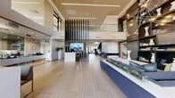 Located at 3140 Saint Rose Parkway in Henderson, Nevada, this elegantly designed lobby lounge delivers a striking first impression with its dramatic double-height ceilings and expansive open floor plan. Large-format tile flooring in a warm neutral tone runs throughout, lending a cohesive and polished feel to the entire space.

The living area is anchored by a sleek, linear gas fireplace set into a textured stone accent wall, flanked by floor-to-ceiling windows that invite abundant natural light and outdoor views. Contemporary blue lounge chairs and sofas are thoughtfully arranged atop patterned area rugs, creating intimate yet open seating vignettes. A bold glass staircase with illuminated wall sconces serves as a striking architectural centerpiece, rising against a contrasting stone feature wall adorned with curated artwork.

A large white tile accent wall provides a clean, gallery-like backdrop to one side of the lounge, while a built-in dark shelving unit displays decorative objects and books. A glass-enclosed workspace, visible through full-height glazing, offers a dedicated area furnished with desks and task chairs. The overall ambiance is modern and upscale, combining rich textures, dramatic lighting, and carefully selected furnishings to create a welcoming yet refined communal environment.