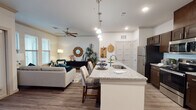 This single-floor residence offers a thoughtfully designed open-concept layout that seamlessly connects the kitchen, dining, and living areas. The kitchen is a functional centerpiece, featuring dark espresso cabinetry, granite countertops, stainless steel appliances, and a large island with an integrated sink and bar seating — ideal for both meal preparation and casual dining. Pendant lighting above the island and recessed ceiling lights provide ample illumination throughout.

The living area flows naturally from the kitchen, offering a bright and welcoming atmosphere enhanced by multiple windows with plantation-style blinds that allow abundant natural light. Crown molding accents the ceiling, and a ceiling fan adds to the comfort of the space. Warm-toned wood-look flooring runs consistently throughout the main living areas.

The bedroom is generously proportioned, providing ample space for a full suite of furniture. A walk-in closet with built-in shelving offers practical storage. The primary bathroom is well-equipped with a dual-sink vanity topped with granite, dark espresso cabinetry, a large mirror, contemporary lighting, and a glass-enclosed walk-in shower. Wood-look flooring continues into the bathroom, maintaining a cohesive aesthetic throughout the residence.