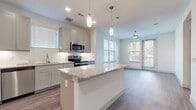 This well-appointed single-floor residence showcases a thoughtful open-concept layout that seamlessly connects the kitchen, dining, and living areas. The kitchen is a standout feature, equipped with stainless steel appliances including a range, microwave, dishwasher, and refrigerator. Gray shaker-style cabinetry lines the walls, complemented by granite countertops and a clean white tile backsplash. A central island with a matching granite surface provides additional workspace and casual seating, while stylish pendant lights hang overhead, adding a modern touch. Rich wood-look flooring runs throughout the main living areas, enhancing the warm, contemporary aesthetic. The living area benefits from multiple windows dressed with horizontal blinds, allowing generous natural light to fill the space, and a ceiling fan ensures comfortable airflow. A sliding glass door leads to an outdoor area, extending the living space further. The bedroom features plush carpeting, a ceiling fan, and a large window that brings in ample daylight. The bathroom is finished with a dual-sink vanity topped with granite, gray cabinetry with brushed gold hardware, dark-framed mirrors, hardwood-style flooring, and a glass-enclosed shower. Throughout, the residence maintains a cohesive, modern palette of neutral tones, creating a clean and inviting atmosphere.