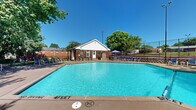 This outdoor recreational space centers around a generously sized swimming pool with crystal-clear turquoise water, graded from a shallow end of 3 feet to a deeper section of 7 feet, making it suitable for a wide range of swimmers. The pool is framed by a broad concrete and paved sundeck that provides ample room for lounging, lined with numerous chairs and seating arrangements positioned to take full advantage of the open sky and abundant natural sunlight.

Multiple patio umbrellas are stationed throughout the deck, offering shaded retreats for relaxation. A brick-and-white-siding clubhouse structure anchors one end of the pool area, featuring a covered entryway, informational signage, and exterior storage units, serving as a functional hub for the amenity space.

Mature trees border the perimeter, lending natural shade, privacy, and a lush, park-like atmosphere to the setting. A fenced tennis court with overhead lighting is situated adjacent to the pool area, adding an additional recreational dimension to the space. The surrounding brick residential buildings are visible beyond the tree line, contextualizing this as a well-appointed community amenity area designed for active outdoor leisure and social gathering under open skies.