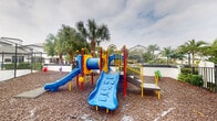 This outdoor playground is a lively, well-equipped recreational space designed for children. The centerpiece is a bold, multi-component play structure finished in primary colors — red, yellow, and blue — incorporating a tunnel, climbing frames, a wide blue slide embossed with ocean-themed motifs such as crabs and starfish, and an additional smaller slide. A separate spring-mounted ride-on toy in green adds further play variety. The entire play area is surfaced with a generous layer of wood chip mulch, providing a safe, impact-absorbing ground cover beneath and around all equipment. The space is enclosed by a dark metal fence, defining its boundaries while maintaining clear sightlines for supervision. Mature tropical palm trees frame the perimeter, lending a lush, verdant backdrop and providing partial shade. Surrounding landscaping includes low shrubs and greenery, contributing to the area's tidy, maintained appearance. Residential buildings are visible beyond the fencing, indicating the playground serves a community setting. The open-air layout allows for ample natural light and airflow, creating an inviting environment for active outdoor play.