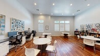 Located at 24011 Richards Road in Spring, TX, this single-floor space is thoughtfully designed to support both collaborative and individual activities. The main open area is the centerpiece of the space, boasting soaring ceilings and recessed lighting that create a bright, welcoming atmosphere. Rich hardwood flooring runs throughout, adding warmth against the crisp white walls. Multiple round tables paired with modern white chairs are arranged throughout the room, offering flexible seating configurations suited for group work or casual gatherings. Natural light filters in through a series of windows fitted with white blinds, positioned near the ceiling line to maximize illumination while maintaining privacy. A built-in workstation along one wall features several computer terminals with ergonomic chairs, well-suited for individual tasks. Bookshelves stocked with books and materials line another wall, accompanied by decorative artwork that adds a creative touch. A compact kitchenette with a sink and dark countertop cabinetry provides practical utility, and a glass-paned interior door connects to an adjacent fitness area, expanding the overall functionality of the space.