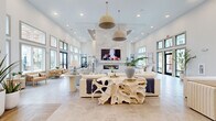 This single-story space is designed in a coastal-modern style, offering a generous, open layout with soaring ceilings and an abundance of natural light streaming through expansive floor-to-ceiling windows. The main lounge area serves as the centerpiece, featuring a dramatic floor-to-ceiling marble fireplace wall with a mounted flat-screen display, woven pendant light fixtures, and sculptural organic-form furnishings atop large-format tile flooring. Multiple seating arrangements invite both social gatherings and quiet relaxation.

Adjacent to the lounge, a dedicated dining and kitchenette area transitions to warm herringbone-patterned wood-look tile flooring. This zone is equipped with rich walnut cabinetry, quartz countertops, a brass pull-down faucet, and a teal penny-tile backsplash that adds a vibrant coastal accent. Open shelving styled with decorative objects provides visual interest, while a sculptural gold cluster pendant overhead adds a bold design statement.

Entry areas feature tall glazed doors with views of outdoor palm-lined grounds and a pool terrace, while cozy reading nooks with leather accent chairs and coastal artwork offer intimate retreats within this bright, well-appointed communal space.
