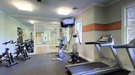 This fitness center offers a comprehensive workout environment designed to meet a variety of exercise needs. The space is finished with durable dark rubber flooring, crisp white crown molding, and a distinctive horizontal orange accent stripe that adds a energetic visual element to the light gray walls. Natural light filters in through a window, complementing the overhead flush-mount lighting to keep the space bright and inviting.

The cardio area is well-stocked with Matrix treadmills, an upright stationary bike, an elliptical trainer, and a stair-stepping machine, providing ample options for aerobic exercise. A wall-mounted flat-screen television is positioned for easy viewing during workouts, enhancing the overall exercise experience. The room also features large mirrors along one wall, creating a sense of expanded space.

French glass panel doors open into an adjacent mirrored room, which appears to offer additional workout or stretching space with views of lush greenery outside through large windows. The overall design is clean, functional, and purpose-built, making this fitness center a practical and well-appointed amenity for residents or users of the facility.