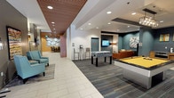 The lobby at 206 Bell St in Seattle, WA offers a welcoming and well-appointed common area designed for both relaxation and social interaction. The space features a contemporary aesthetic, highlighted by a distinctive slatted wood ceiling that adds warmth and texture, complemented by recessed lighting and clean white tile flooring that transitions into patterned carpet in the activity zones.

The game and recreation area serves as a central focal point, equipped with a full-size pool table and a foosball table, making it an ideal gathering spot for residents or guests. Adjacent to the game area is a comfortable lounge zone furnished with an orange sofa, wall-mounted television, and curated wall art, creating an inviting atmosphere for casual socializing.

A dedicated seating nook features bold textured orange wallcovering, a curved sectional sofa, and coordinating artwork, offering a more intimate setting for smaller gatherings or quiet relaxation. The main corridor area incorporates teal accent chairs and decorative wall installations, adding pops of color throughout. A reception counter area and elevator access are also visible, along with glass entry doors that allow natural light to fill the space. The overall design blends modern finishes with thoughtful amenity offerings, creating a functional and visually engaging lobby environment.