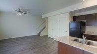 This two-story space is designed with an open and functional layout, beginning on the main floor where the living and kitchen areas flow seamlessly together. The flooring throughout the main level consists of durable hardwood-style planks in a warm, medium-toned finish, lending a clean and cohesive look to the space. A ceiling fan is installed in the living area, providing both air circulation and a subtle decorative element overhead.

The kitchen is positioned adjacent to the living space and features a peninsula or counter with a granite-style countertop in neutral tones, complemented by warm wood-toned cabinetry. A black refrigerator anchors the kitchen area, and the open sightlines between the kitchen and living room enhance the sense of spaciousness on the main floor.

A staircase with white-painted railings and balusters leads to the upper level, adding a classic, tidy architectural element. Nearby, a set of double closet doors with paneled detailing provide convenient storage on the main floor. The walls are painted in a soft, neutral white throughout, keeping the interior bright and versatile. Overall, the space presents a practical and well-organized layout suited to everyday living.
