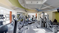 This fitness center offers a bright, energetic environment designed to support a full range of workout routines. The space is outfitted with an array of cardio equipment, including treadmills, elliptical machines, and stationary bikes, all arranged to allow comfortable circulation throughout the floor. A multi-station cable and strength training machine is positioned along one side, providing options for resistance and functional training.

The interior design is notably contemporary, featuring a striking ceiling adorned with sweeping geometric panels in shades of yellow-green and blue, adding a dynamic visual element overhead. The flooring consists of patterned carpet in neutral tones with subtle linear accents, contributing to the modern aesthetic while providing a comfortable surface underfoot.

Large windows along one wall allow natural light to fill the space, creating an open and inviting atmosphere. A mirrored wall on the opposite side visually expands the room and allows users to monitor their form during workouts. The overall ambiance is clean, well-lit, and thoughtfully designed to motivate and accommodate a variety of fitness activities.