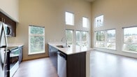 This thoughtfully designed single-floor residence combines modern aesthetics with practical living. The open-concept living and kitchen area is anchored by dramatically high ceilings and an abundance of natural light streaming through expansive multi-pane windows, offering serene views of surrounding evergreen trees and the neighboring community. The kitchen features a sleek center island with a white quartz countertop, an undermount sink, dark espresso cabinetry, and a full suite of stainless steel appliances including a refrigerator, range, and microwave. Hardwood-style flooring flows consistently throughout the space, complementing the warm neutral tones on the walls. The living area is generously proportioned, with a glass-panel door leading to a private balcony. The bedroom continues the airy feel with soaring ceilings and dual windows framing tree-lined views. A well-appointed full bathroom includes a dark-toned vanity with a quartz countertop and a full tub-shower combination. A dedicated laundry area houses a stacked washer and dryer unit, adding everyday convenience. A walk-in closet with built-in shelving completes the bedroom suite. The overall ambiance is clean, bright, and contemporary throughout.