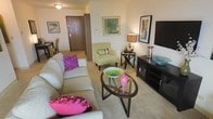 This single-floor residence offers a thoughtfully designed living area that serves as the heart of the space. The living room is furnished with a generously sized neutral-toned sofa accented by bold purple throw pillows, complemented by a light green accent chair that adds a refreshing pop of color. A sleek oval glass coffee table anchors the seating area, topped with decorative bowls and vases in vivid pink and green hues that bring energy to the neutral palette.

The entertainment area features a dark wood media console housing a flat-screen television, with decorative accessories adding visual interest. A warm-toned table lamp provides ambient lighting near the accent chair, contributing to the room's cozy atmosphere. Wall art in coordinating tones is tastefully displayed throughout, enhancing the curated feel of the space.

Plush wall-to-wall carpeting in a warm beige tone runs throughout, adding comfort underfoot. A glimpse of a dining area is visible toward the rear of the space, suggesting a functional open layout. The solid wood entry door with its distinctive panel design provides a grounded, classic touch to the overall aesthetic.