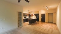 This single-floor space showcases a thoughtfully designed open-concept layout that integrates the living area and kitchen into one cohesive environment. The kitchen is a standout feature, outfitted with dark espresso cabinetry, black appliances including a refrigerator, stove, and microwave, and a central island that provides additional prep and seating space. A pendant light hangs above the island, complementing the track lighting that runs along the ceiling to illuminate the kitchen workspace effectively.

The flooring transitions from light hardwood in the kitchen and entry areas to plush carpet in the living space, creating a subtle yet functional distinction between zones. A built-in open shelving unit is recessed into the wall near the living area, offering convenient display or storage space. The entry door area is clearly defined, providing a natural transition point into the unit.

The ceiling features a ceiling fan in the living area, adding both functionality and a modern touch. The overall color palette — warm neutrals paired with dark cabinetry — lends the space a contemporary yet welcoming ambiance, making it feel both stylish and practical for everyday living.