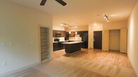 This single-floor space showcases a thoughtfully designed open-concept layout that integrates the living area and kitchen into one cohesive environment. The kitchen is a standout feature, outfitted with dark espresso cabinetry, black appliances including a refrigerator, stove, and microwave, and a central island that provides additional prep and seating space. A pendant light hangs above the island, complementing the track lighting that runs along the ceiling to illuminate the kitchen workspace effectively.

The flooring transitions from light hardwood in the kitchen and entry areas to plush carpet in the living space, creating a subtle yet functional distinction between zones. A built-in open shelving unit is recessed into the wall near the living area, offering convenient display or storage space. The entry door area is clearly defined, providing a natural transition point into the unit.

The ceiling features a ceiling fan in the living area, adding both functionality and a modern touch. The overall color palette — warm neutrals paired with dark cabinetry — lends the space a contemporary yet welcoming ambiance, making it feel both stylish and practical for everyday living.
