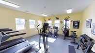 This fitness center offers a welcoming and energetic environment designed to support a well-rounded workout routine. The space is bathed in natural light streaming through multiple windows along the walls, creating an open and motivating atmosphere. The walls are painted in a warm yellow hue and are accented with colorful, playful pop-art style murals, adding a fun and vibrant character to the room. The flooring consists of durable, easy-to-clean tile and rubberized surfaces, practical for high-traffic exercise use. The center is equipped with a solid selection of cardio machines, including a treadmill, elliptical trainer, and stationary bike, catering to a range of cardiovascular fitness goals. Strength training is also well-represented, with a multi-function cable and weight machine available for resistance-based workouts. Overhead fluorescent lighting supplements the natural light, ensuring the space remains well-lit at all times of day. The layout is thoughtfully arranged to allow comfortable movement between equipment. Overall, this fitness center combines functionality with an upbeat aesthetic, making it an inviting destination for residents or members looking to maintain an active lifestyle.