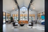 Located at 240 Martin Rd SW in Huntsville, Alabama, the Belmont Hill leasing office is a single-story space designed to make a strong first impression. The open-concept layout is defined by dramatic vaulted ceilings and bold dark structural columns that contrast sharply against crisp white walls, creating a modern yet welcoming atmosphere. Large multi-pane windows line the perimeter, flooding the interior with natural light and offering views of the surrounding community, including the outdoor pool area. The main reception and lounge area features stylish contemporary furnishings, including a tufted amber sofa, velvet accent chairs in soft gray, and a round wood coffee table — all arranged on a patterned area rug. Geometric gold pendant lights hang from the ceiling, adding warmth to the space. A dedicated workstation area is equipped with a desk, seating for guests, and a wall-mounted television, making it functional for leasing consultations. Near the entrance, additional seating provides a transitional welcome zone. French-style interior doors and light wood-tone flooring throughout contribute to a cohesive, polished aesthetic. The overall ambiance balances professional functionality with inviting residential comfort, reflecting the character of the Belmont Hill community.
