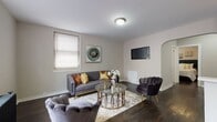 This well-appointed single-floor residence offers a thoughtfully designed layout that balances comfort and modern style. The living room serves as the heart of the space, featuring rich dark hardwood flooring, neutral warm-toned walls, and multiple windows fitted with blinds that allow for controlled natural light. A cast-iron radiator adds a classic architectural touch, complementing the room's contemporary furnishings and arched entryway detail.

The kitchen is bright and functional, outfitted with crisp white shaker cabinetry, light wood-tone flooring, and a full suite of stainless steel appliances including a gas range, over-the-range microwave, and refrigerator. A peninsula provides additional counter space and a natural transition into the adjacent dining area, which comfortably accommodates a small dining set.

The bedroom is serene and well-proportioned, with the same dark hardwood flooring found throughout the living areas. It includes a dedicated closet and is connected to a hallway that also features an additional storage closet.

The bathroom is finished with marble-look wall tile surrounding the soaking tub and shower, a window for natural ventilation, and a classic cast-iron radiator that echoes the vintage character found elsewhere in the space. Clean white fixtures complete the polished look.
