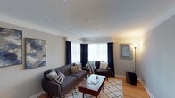 This single-floor space showcases a thoughtfully designed living room that balances comfort with style. The room features smooth, white ceilings with recessed lighting that provides even illumination throughout the space. Neutral grey walls create a versatile backdrop, complemented by warm hardwood flooring that adds natural character to the interior.

A bay window serves as a focal point, dressed with elegant navy blue curtains framing sheer white panels that allow natural light to filter softly into the room. The seating arrangement centers around a tufted charcoal grey sofa adorned with decorative pillows, paired with a matching dark armchair, both positioned around a rich wooden coffee table. A geometric-patterned area rug in white and grey anchors the seating area, adding texture and visual interest to the space.

Decorative accents include an abstract blue and gold wall painting, a silver ornate mirror, and a tall arc floor lamp that adds both function and elegance. A woven basket in the corner provides practical storage while enhancing the room's warm, curated aesthetic. Overall, the space presents a cohesive, contemporary design with a welcoming atmosphere.