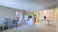 This single-floor residential unit offers a practical and comfortable open-concept layout that seamlessly connects the living area to the kitchen. The living space is finished with plush wall-to-wall carpeting in a neutral tone, creating a warm and inviting atmosphere. Soft, taupe-colored walls complement the light palette throughout, lending the space a clean and cohesive feel.

The kitchen is efficiently designed with light maple cabinetry providing ample storage, black appliances including a refrigerator and range, and a breakfast bar counter that doubles as a casual dining and prep area. The layout ensures easy flow between cooking and living spaces.

A textured ceiling adds visual interest overhead, while the open doorway leads to additional rooms beyond, hinting at a thoughtfully arranged floor plan. The unit benefits from recessed lighting in the kitchen area and natural-feeling ambient light throughout.

Overall, the space strikes a balance between functionality and comfort, with its neutral finishes and open design making it easy to personalize. The cohesive color scheme, practical kitchen features, and open living area create an accessible and livable environment well-suited for everyday life.